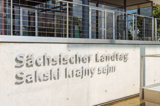 Seit dem Jahr 2018 ist der Besuchereingang des Sächsischen Landtags auch auf Sorbisch beschriftet: „Sakski krajny sejm“. Im Sitzungssaal des Präsidiums steht zudem die Flagge der Sorben. - Bild: Sächsischer Landtag